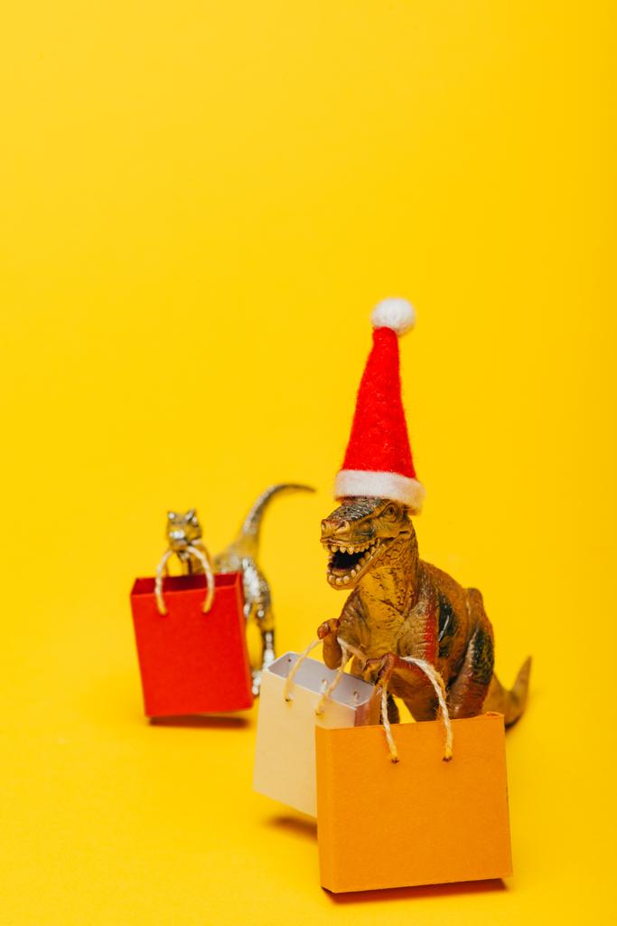 Concentration sélective de dinosaures jouets dans un chapeau de Père Noël et des sacs à provisions sur fond jaune
 - Photo, image
