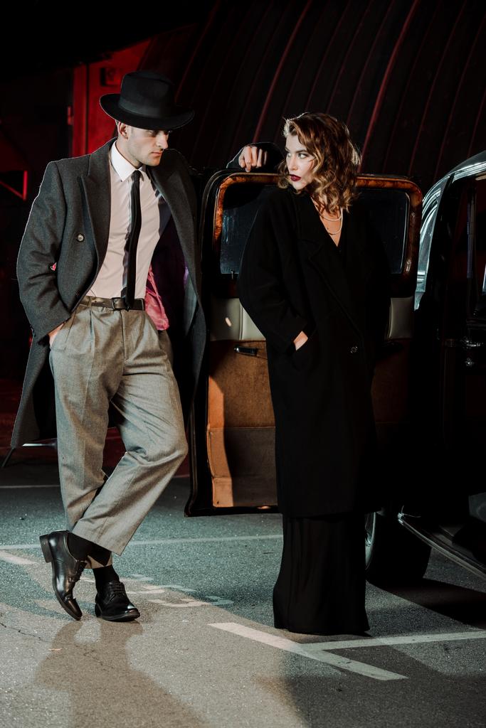 schöner Gangster, der mit der Hand in der Tasche steht und attraktive Frau in der Nähe von Auto ansieht  - Foto, Bild