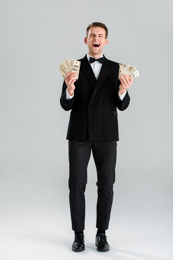 šťastný muž v obleku drží dolarové bankovky na šedé  - Fotografie, Obrázek