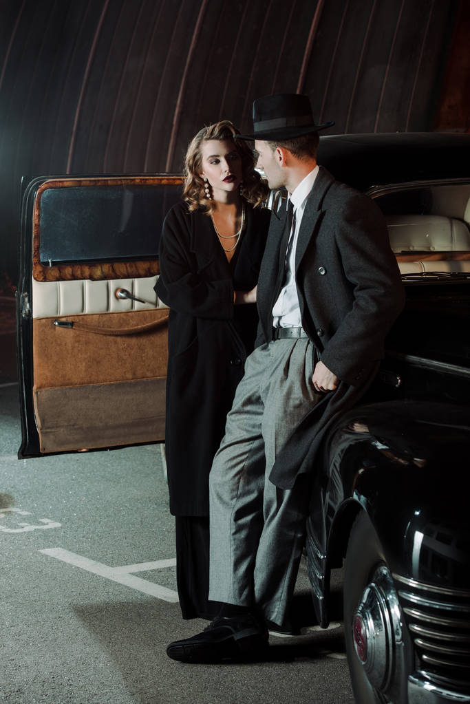όμορφος γκάνγκστερ στο καπέλο κοιτάζοντας ελκυστική γυναίκα κοντά ρετρό αυτοκίνητο  - Φωτογραφία, εικόνα
