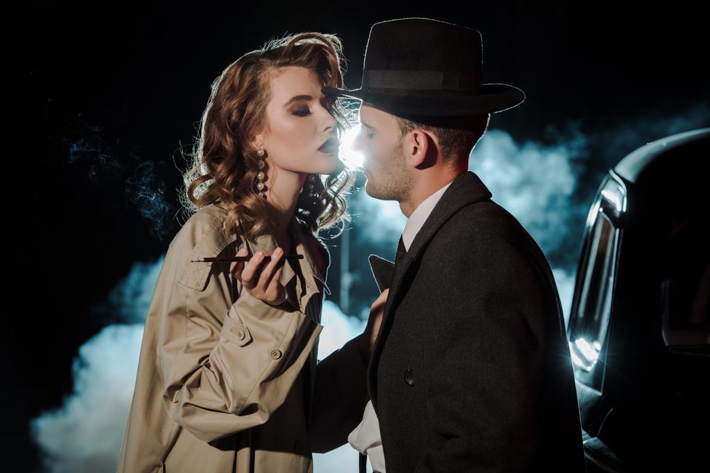 gangster bonito em chapéu olhando para mulher atraente perto de carro em preto com fumaça
  - Foto, Imagem