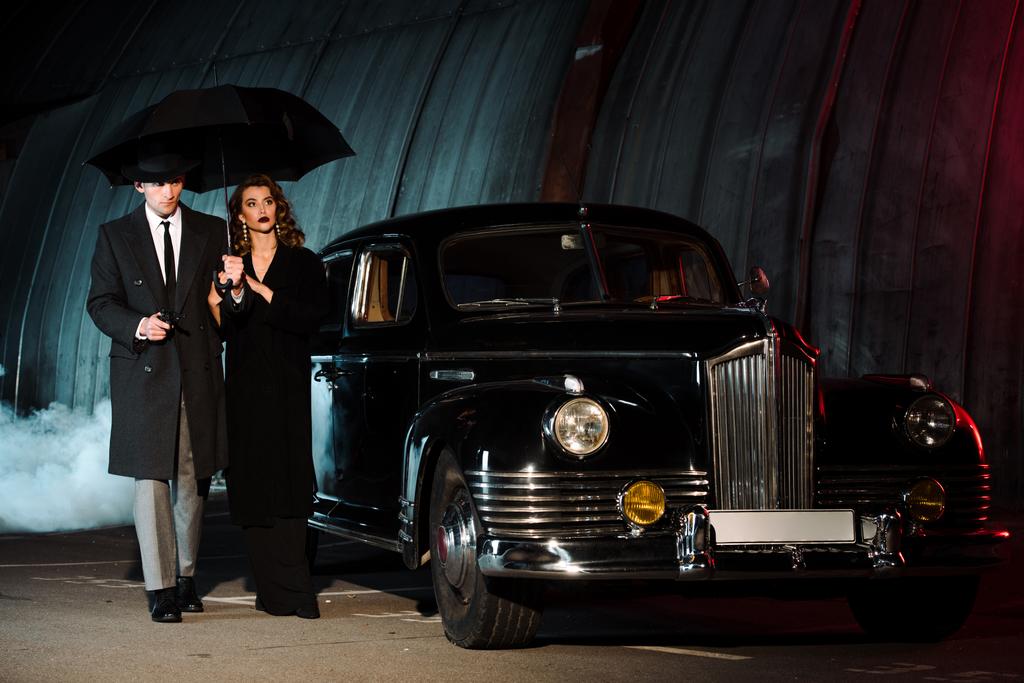przystojny gangster gospodarstwa parasol i pistolet podczas spaceru z atrakcyjną kobietą w pobliżu samochód retro  - Zdjęcie, obraz