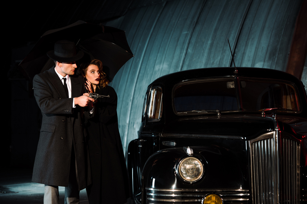 hombre peligroso en sombrero sosteniendo pistola y paraguas cerca de la mujer y coche retro
  - Foto, imagen