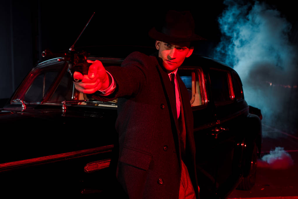 selektywne skupienie stylowy człowiek w kapeluszu trzyma pistolet w pobliżu samochodu retro na czarno z dymem  - Zdjęcie, obraz