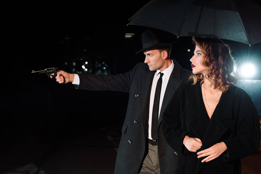 boční pohled na nebezpečného muže v klobouku drží zbraň v blízkosti atraktivní dívka a retro auto na černé - Fotografie, Obrázek