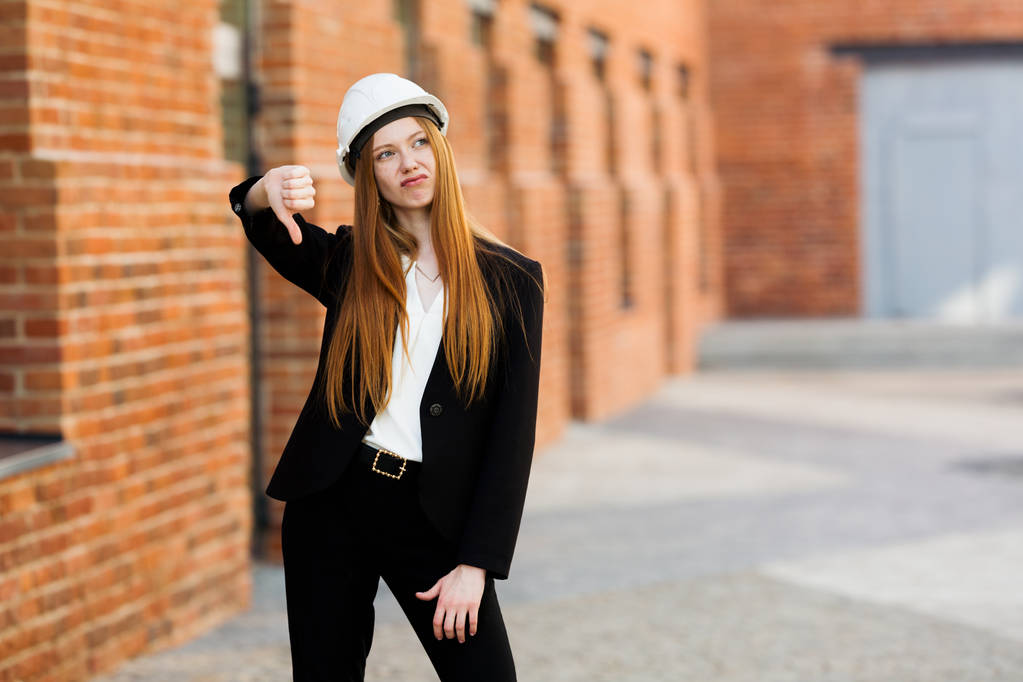 Een roodharig architect meisje in een beschermende helm staat tegen de achtergrond van een bakstenen gebouw en geeft een hekel aan een duim naar beneden als gevolg van mislukking - Foto, afbeelding