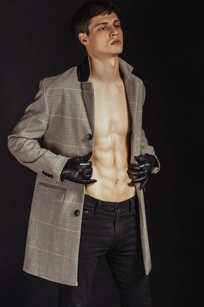 Μύες ισχυρό όμορφο απογυμνωμένο αρσενικό μοντέλο με μαύρα γάντια και παλτό σε τζιν τζιν σε μαύρο απομονωμένο φόντο γραμματοσειράς - Φωτογραφία, εικόνα