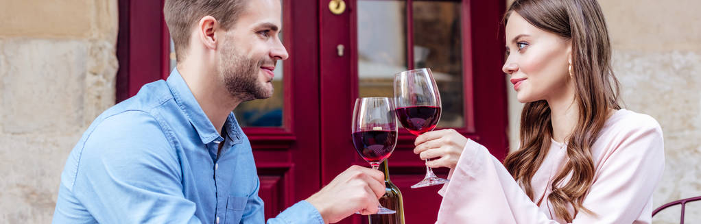 panoramisch schot van glimlachend paar zitten in de straat cafe en klinkende glazen rode wijn - Foto, afbeelding