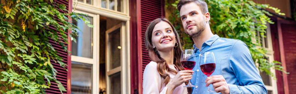Yeşil sarmaşıkla kaplı evin yanında dururken ellerinde kırmızı şarap kadehleri olan mutlu çiftin panoramik görüntüsü. - Fotoğraf, Görsel