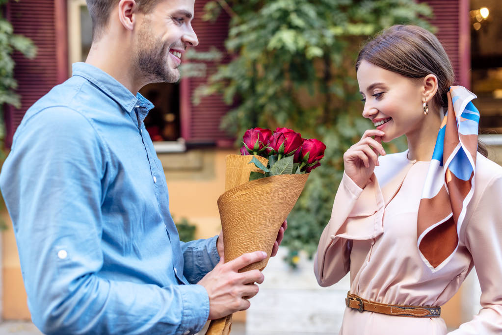 schöner junger Mann überreicht Strauß roter Rosen an glückliche Freundin - Foto, Bild