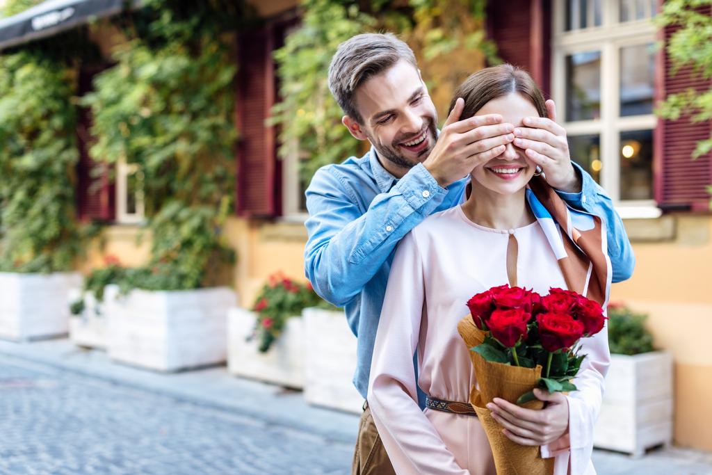 šťastný muž zavírající oči usměvavé přítelkyně držící kytici růží - Fotografie, Obrázek