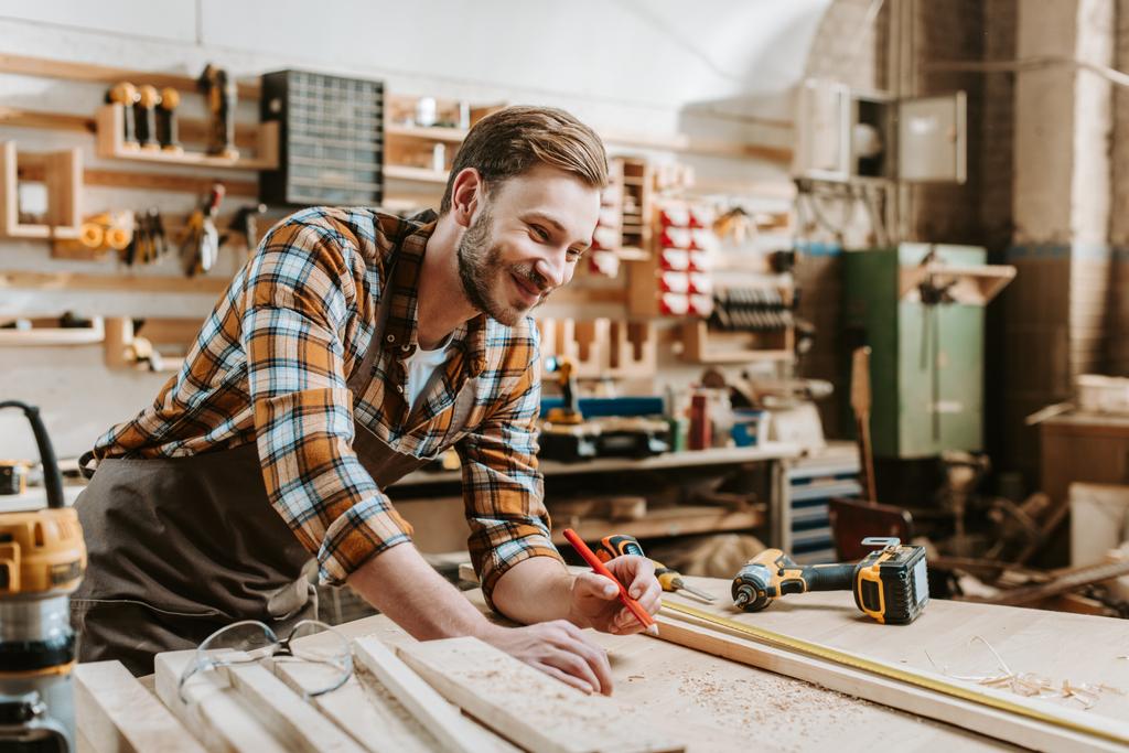 enfoque selectivo de carpintero feliz sosteniendo lápiz cerca de tablón de madera y cinta métrica
  - Foto, Imagen