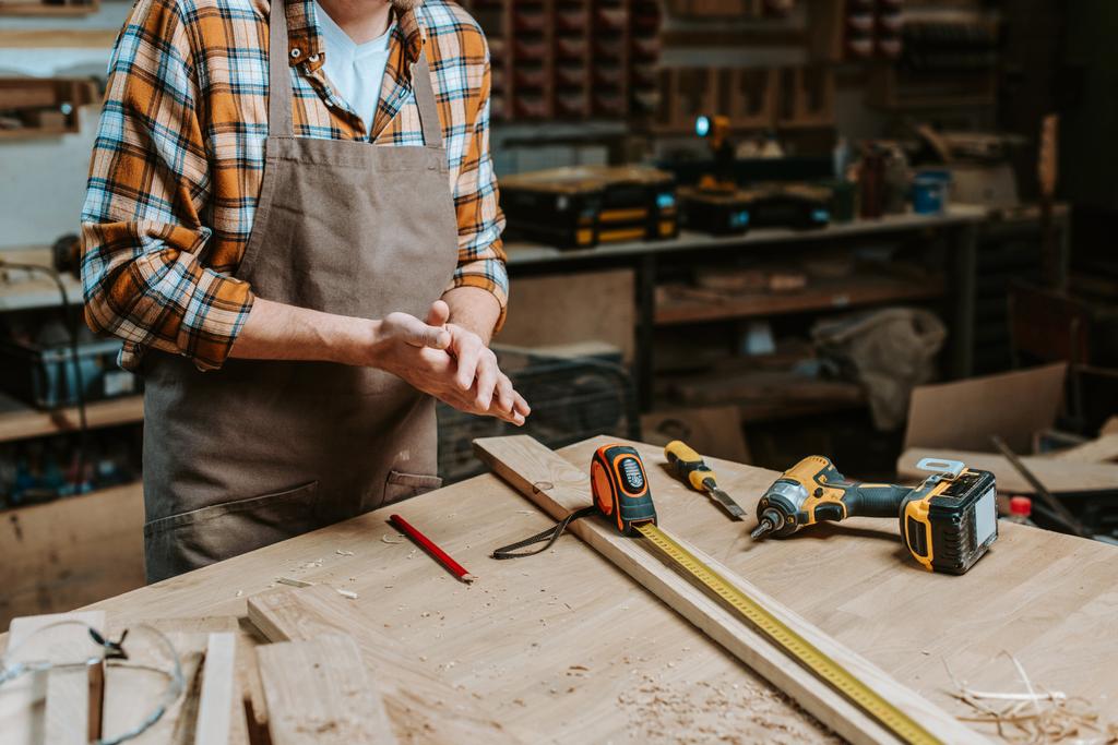 vista recortada de carpintero en delantal de pie con las manos apretadas cerca de la mesa con herramientas
  - Foto, Imagen