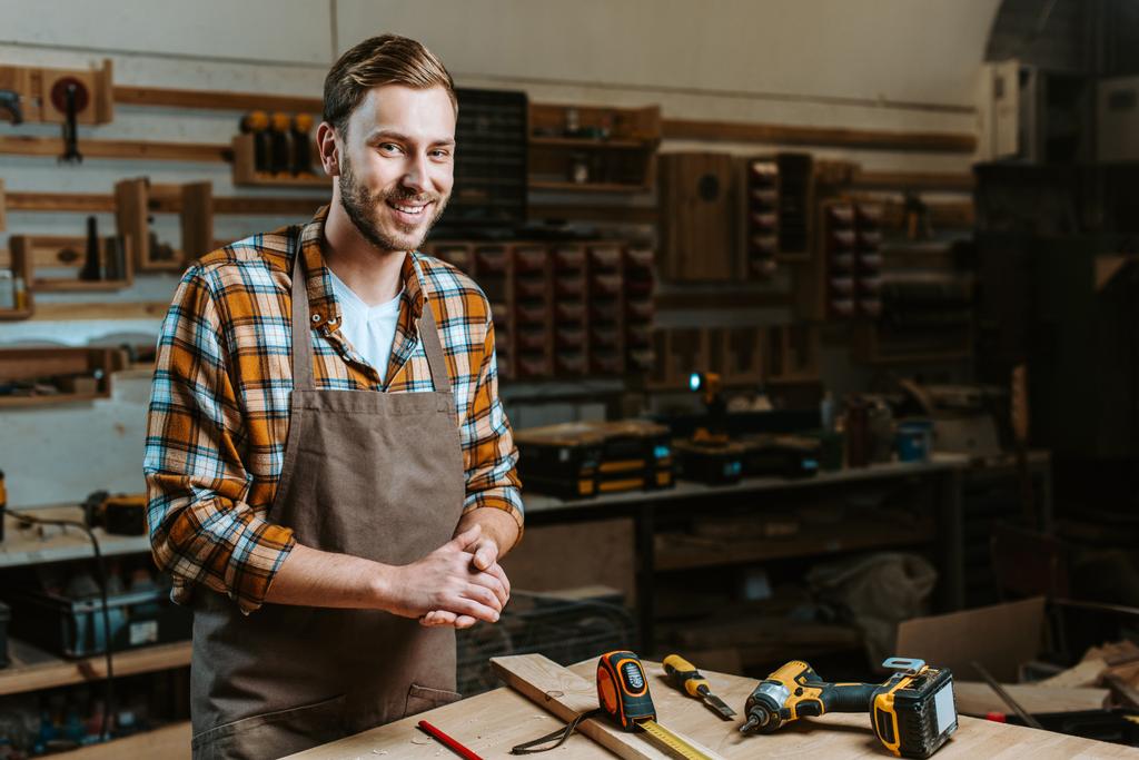 χαρούμενος ξυλουργός με σφιγμένα χέρια στέκεται κοντά στο τραπέζι με εργαλεία  - Φωτογραφία, εικόνα