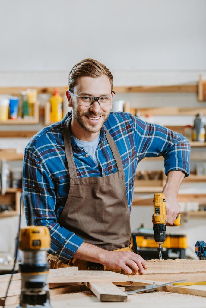 selective focus of cheerful carpenter in goggles and apron holding hammer drill near wooden planks - Photo, Image