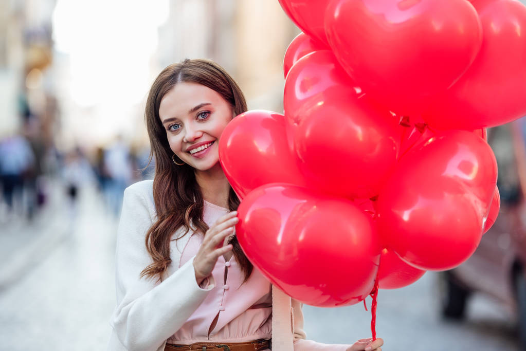 beautiful girl smiling at camera while holding bundle of red heart-shaped balloons - Photo, Image