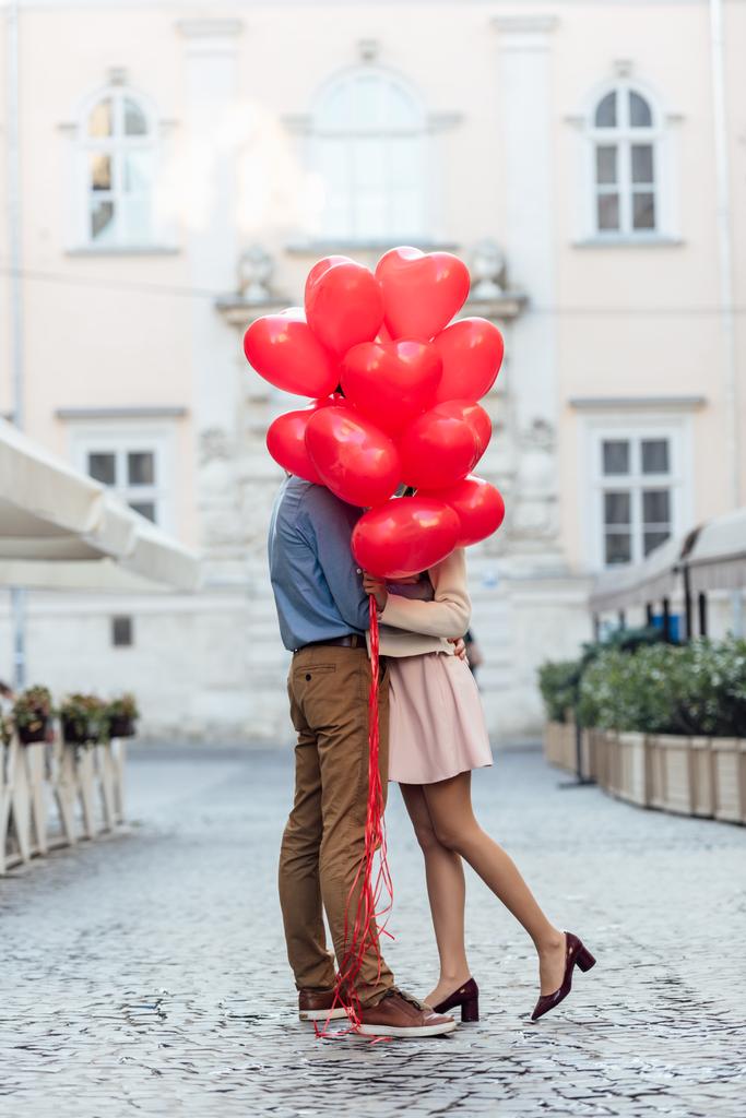 szczęśliwa młoda para obejmująca się chowając się za czerwonymi balonami w kształcie serca na rynku - Zdjęcie, obraz