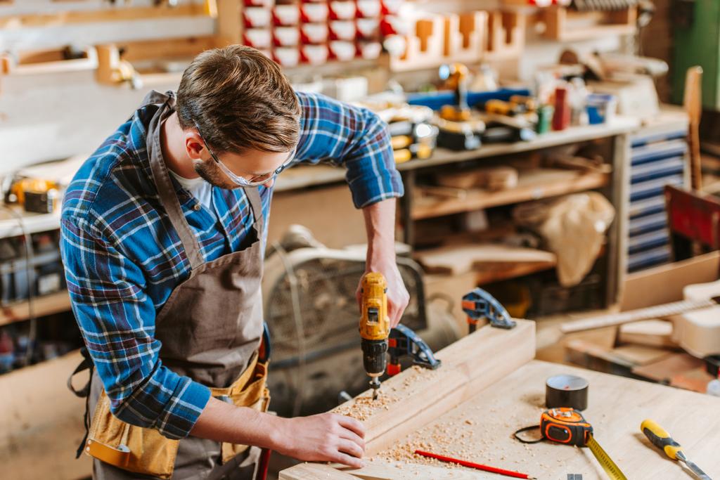 woodworker in goggles and apron holding hammer drill near wooden planks - Photo, Image