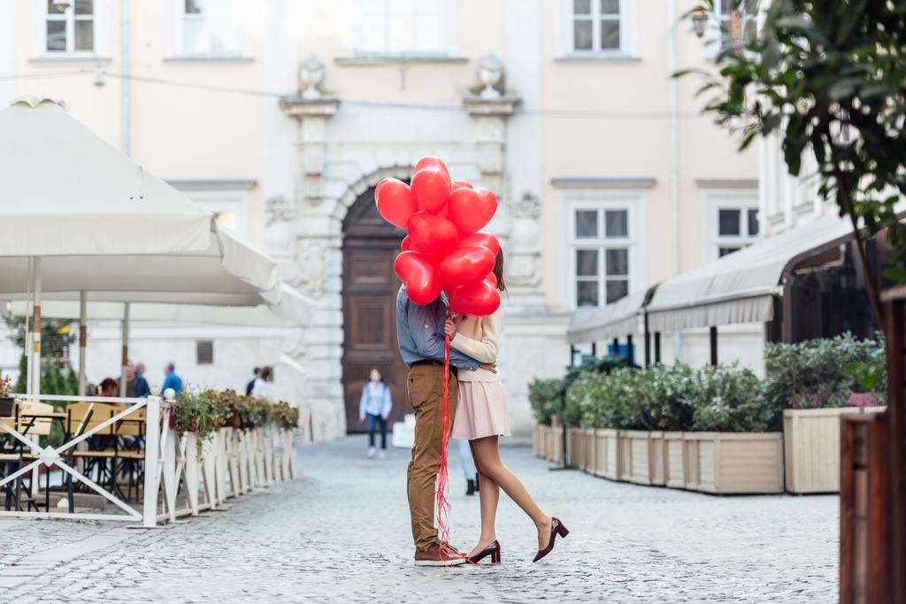 ευτυχισμένο ζευγάρι αγκαλιάζει ενώ κρύβονται πίσω από κόκκινα μπαλόνια σε σχήμα καρδιάς στην πλατεία της πόλης - Φωτογραφία, εικόνα
