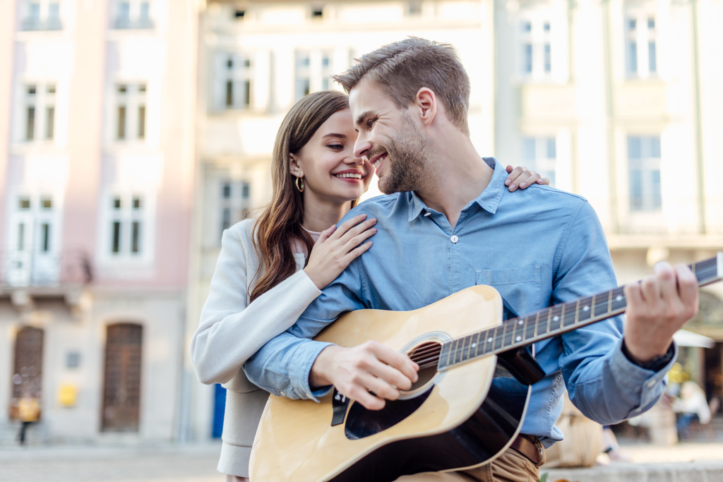 przystojny mężczyzna gra na gitarze akustycznej w pobliżu szczęśliwy dziewczyna na ulicy - Zdjęcie, obraz
