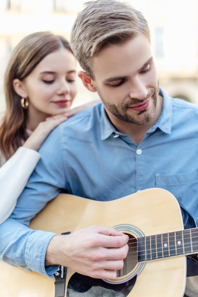 selektivní zaměření pohledný muž hraje na akustickou kytaru poblíž šťastný přítelkyně - Fotografie, Obrázek