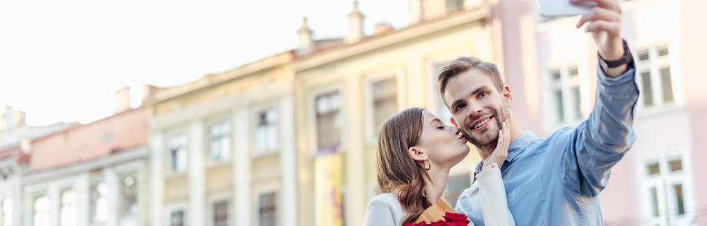 Panoramaaufnahme einer jungen Frau, die ihren Freund küsst und ein Selfie mit dem Smartphone macht - Foto, Bild
