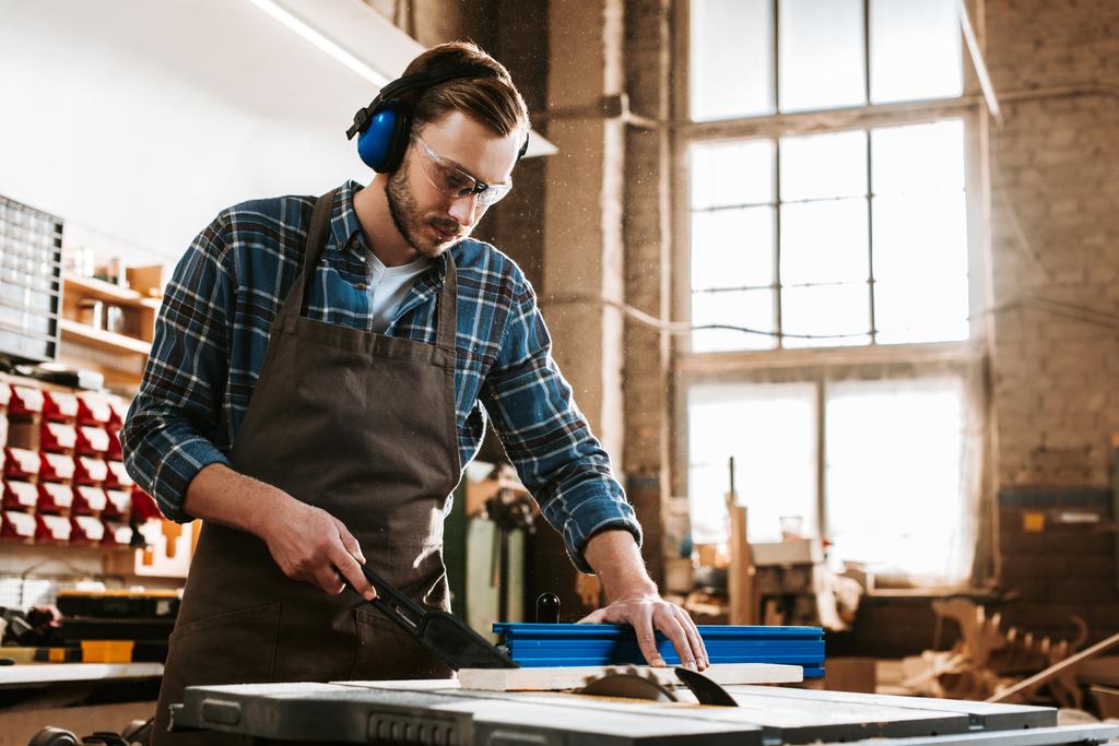 handsome woodworker in protective headphones and apron holding plank near circular saw - Photo, Image