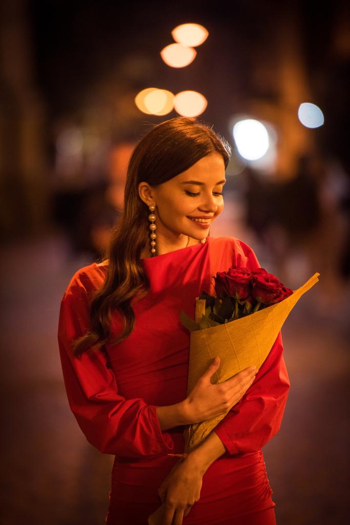 mooi, glimlachend meisje in parel oorbellen met boeket van rode rozen op straat 's nachts - Foto, afbeelding
