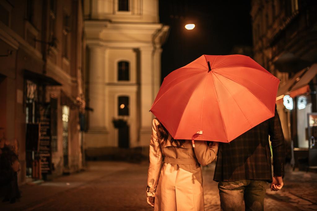 back view of couple in autumn outfit walking under umbrella along evening street - Photo, Image