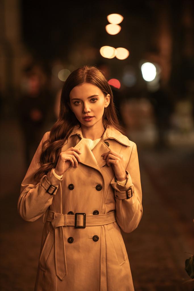 приваблива, впевнена жінка в траншеї пальто дивиться на камеру, стоячи на вечірній вулиці
 - Фото, зображення