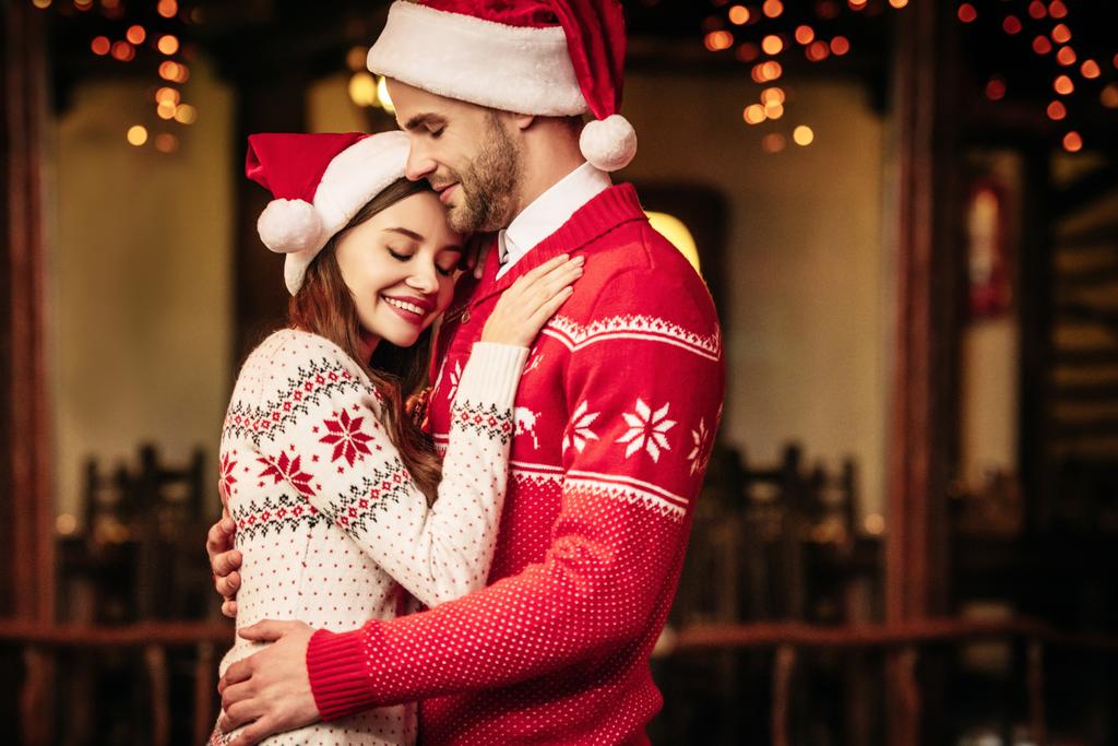 glückliches junges Paar in Weihnachtsmützen und warmen Pullovern, das sich mit geschlossenen Augen umarmt - Foto, Bild