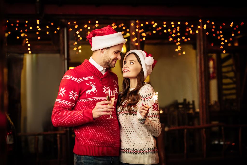 Noel Baba şapkalı mutlu çift ellerinde şampanya bardağı ve maytap tutarken birbirlerine bakıyorlar.  - Fotoğraf, Görsel