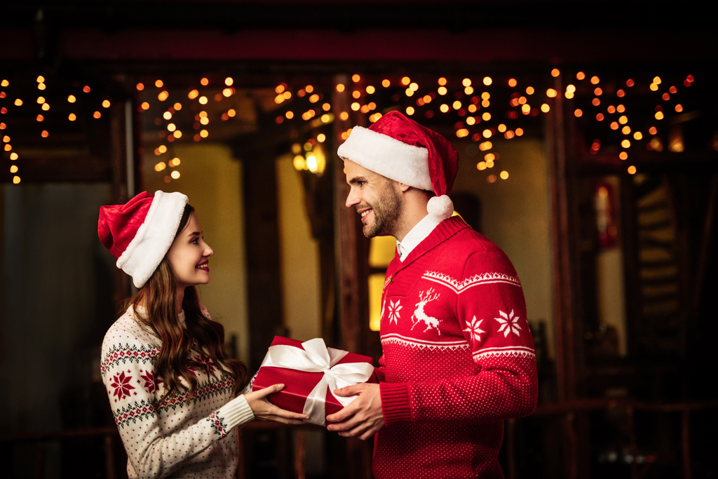 felice giovane coppia in maglioni caldi e cappelli di Babbo Natale in possesso di scatola regalo e guardarsi l'un l'altro
 - Foto, immagini
