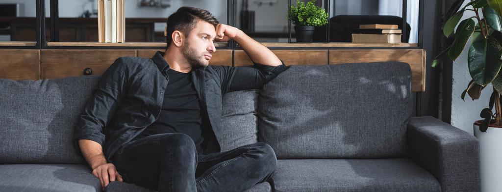 Panoramaaufnahme eines gutaussehenden und nachdenklichen Mannes, der auf einem Sofa in einer Wohnung sitzt  - Foto, Bild