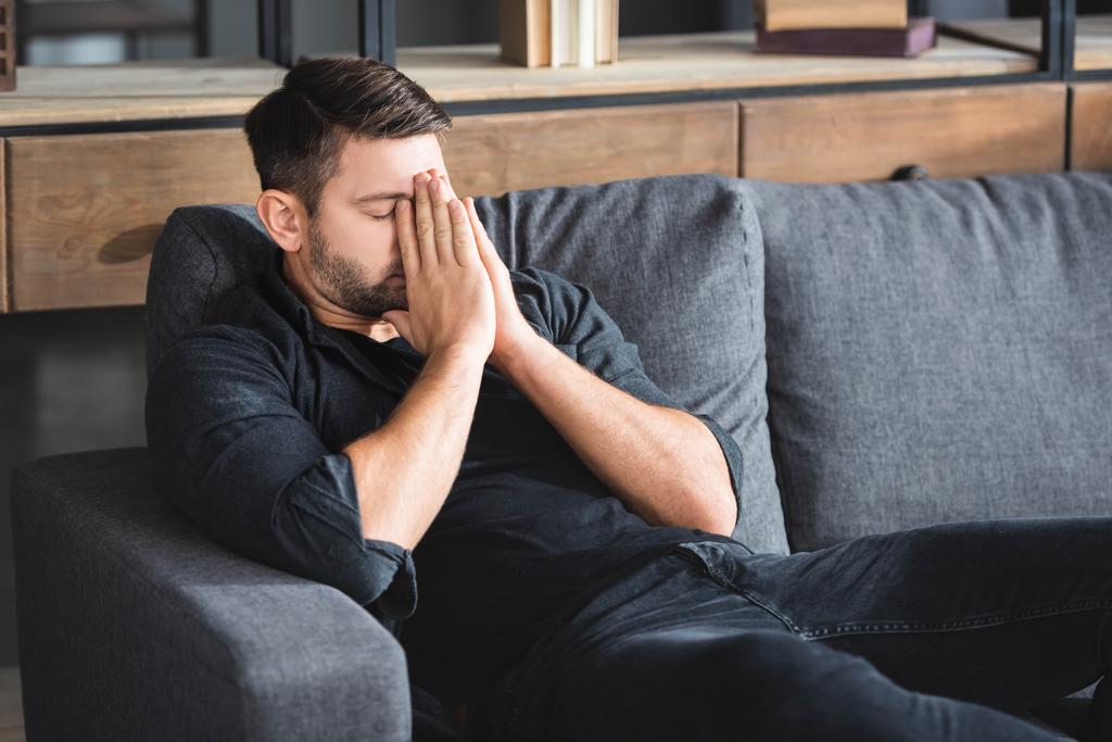 hombre con dolor de cabeza sentado en el sofá y obscurecer la cara en el apartamento
  - Foto, Imagen