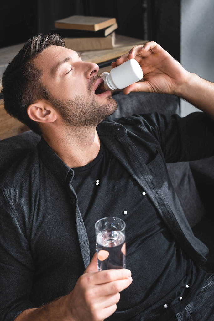 высокий угол зрения красивый мужчина с панической атаки принимая таблетки и держа стакан воды в квартире
  - Фото, изображение