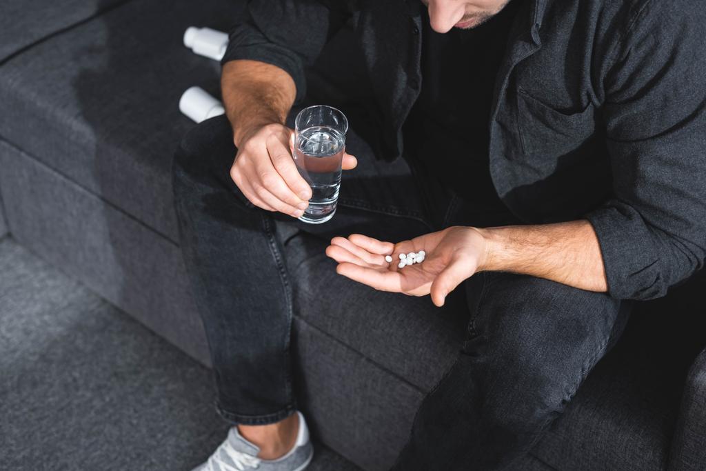oříznutý pohled na člověka s panickým záchvatem držení pilulek a sklenice vody v bytě  - Fotografie, Obrázek
