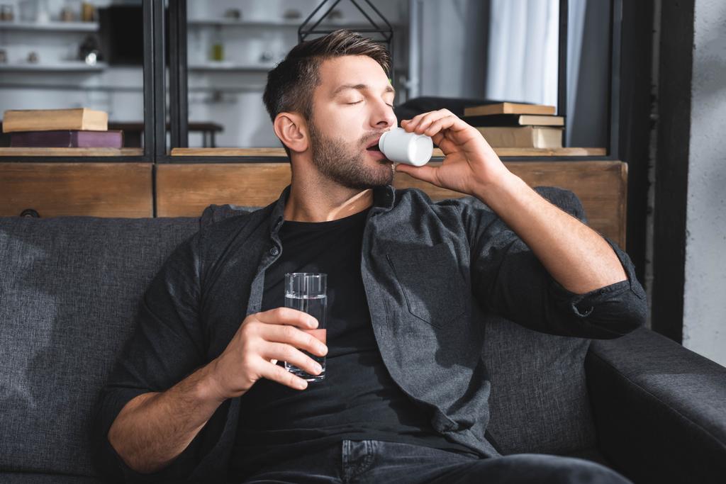 handsome man with panic attack taking pills and holding glass of water in apartment  - Photo, Image