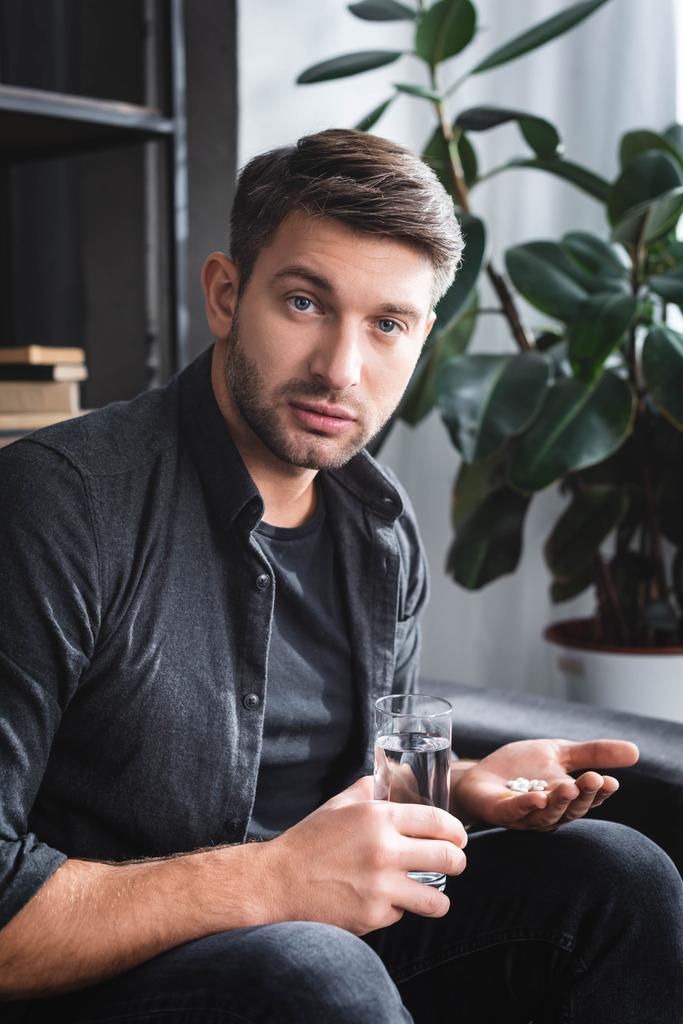 hombre guapo con ataque de pánico sosteniendo pastillas y vaso de agua en el apartamento
  - Foto, Imagen