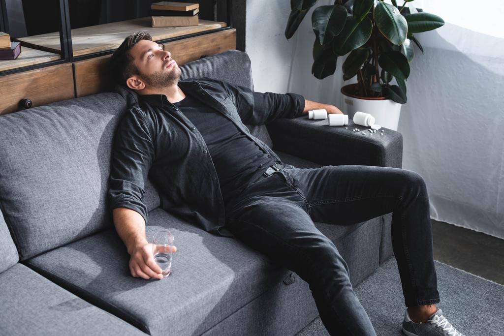 vista de ángulo alto del hombre guapo con ataque de pánico sosteniendo un vaso de agua y sentado en el sofá en el apartamento
  - Foto, imagen