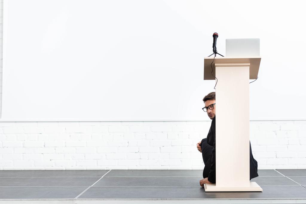 empresário assustado em óculos escondidos atrás tribuno pódio durante conferência isolada em branco
  - Foto, Imagem