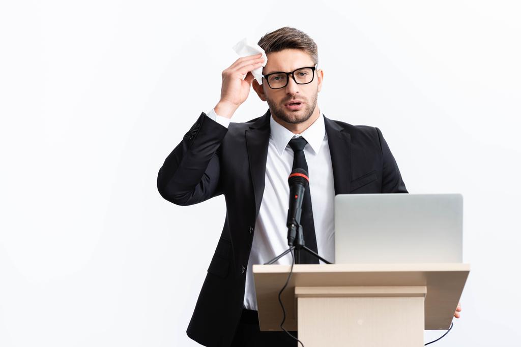 испуганный бизнесмен в костюме стоит на трибуне и держит салфетку во время конференции изолированной на белом
  - Фото, изображение