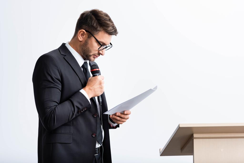 hombre de negocios en traje sosteniendo el micrófono y mirando el papel durante la conferencia aislado en blanco
  - Foto, imagen