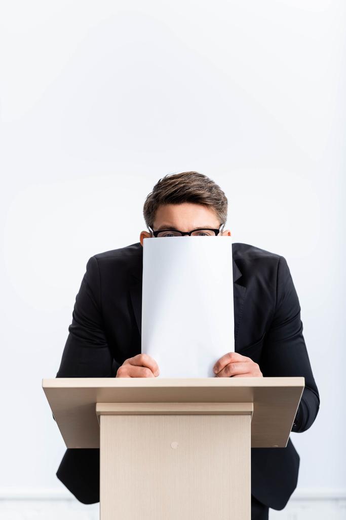 scared businessman in suit standing at podium tribune and obscuring face with paper during conference isolated on white  - Photo, Image