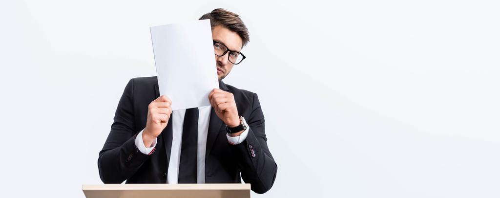 panoramic shot of scared businessman in suit standing at podium tribune and obscuring face with paper during conference  isolated on white  - Photo, Image