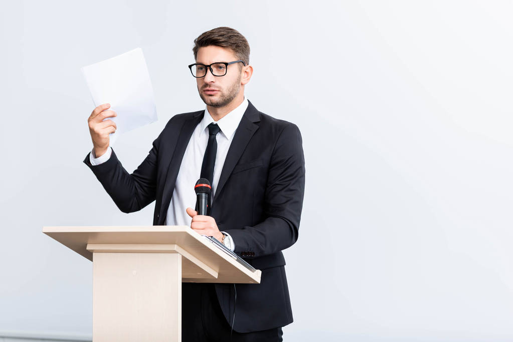 испуганный бизнесмен в костюме стоит на трибуне и держит микрофон во время конференции изолированы на белом
  - Фото, изображение