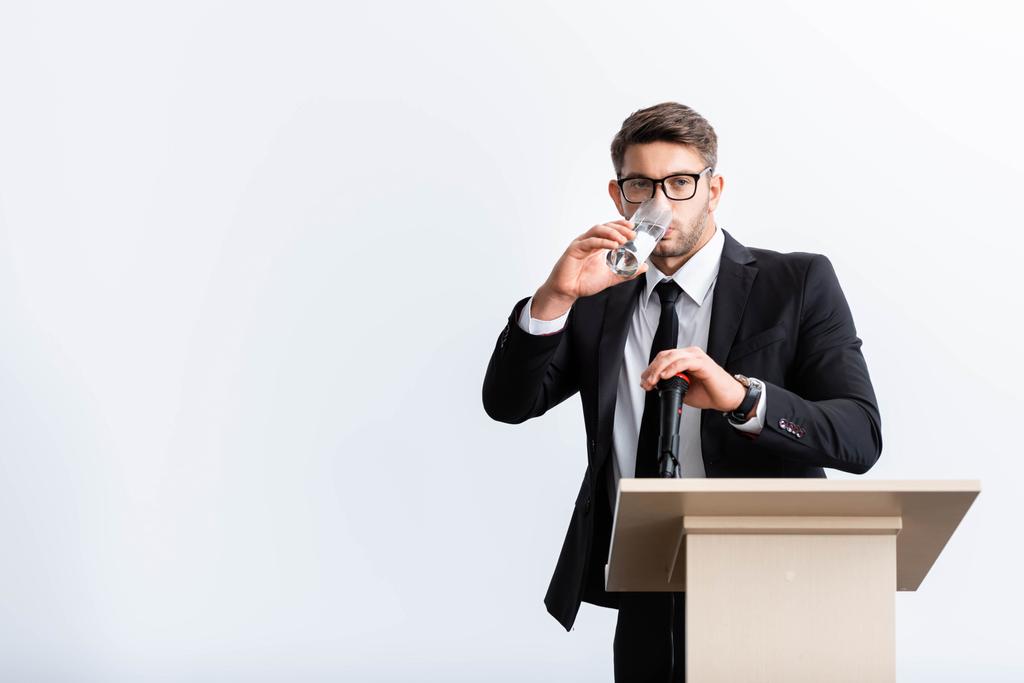 scared businessman in suit standing at podium tribune and drinking water during conference isolated on white  - 写真・画像