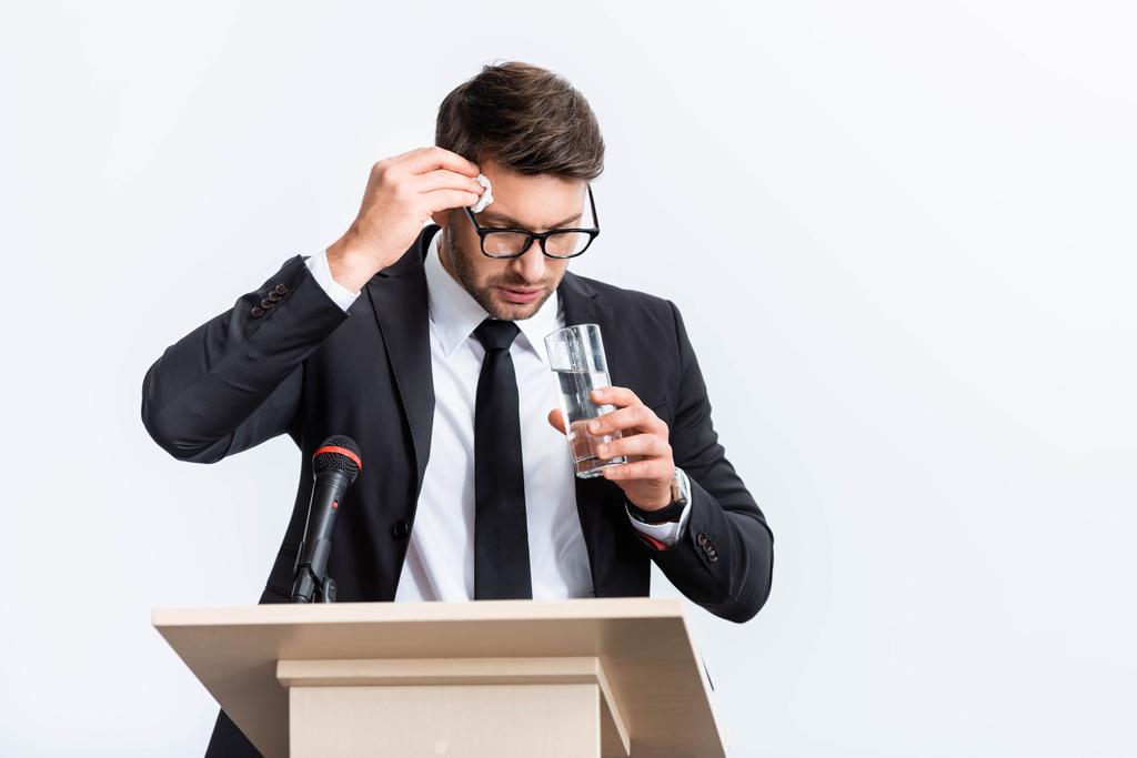 ängstlicher Geschäftsmann im Anzug steht auf der Tribüne und hält während der Konferenz ein Glas Wasser isoliert auf weiß  - Foto, Bild