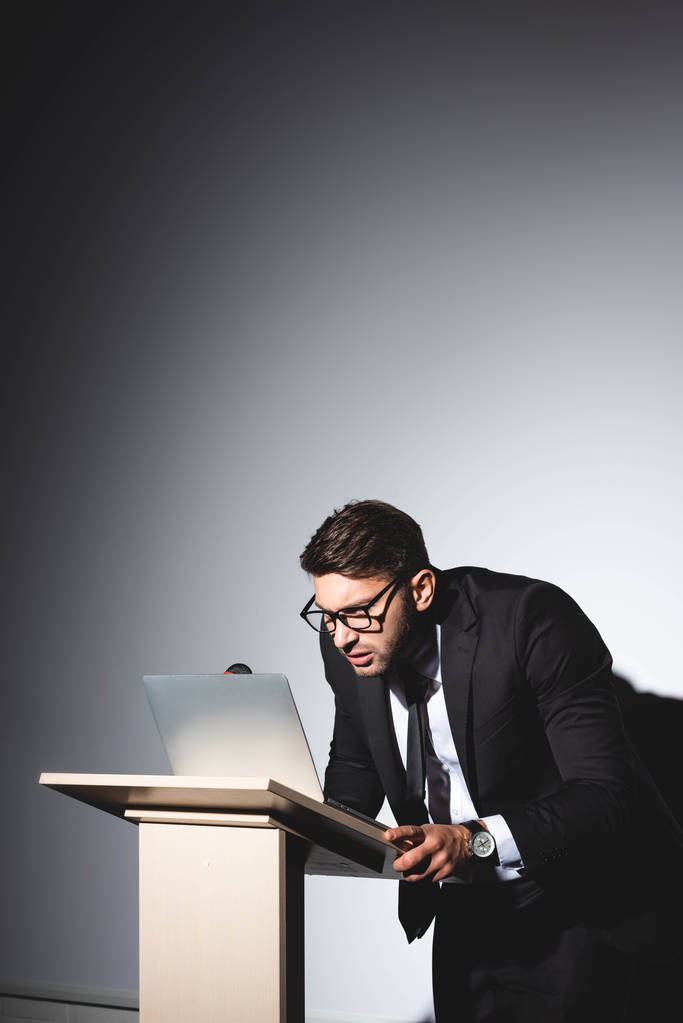 ängstlicher Geschäftsmann im Anzug steht auf der Tribüne des Podiums und schaut auf Laptop während der Konferenz auf weißem Hintergrund  - Foto, Bild