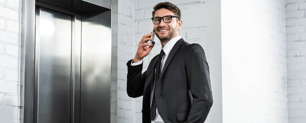 plano panorámico de hombre de negocios sonriente en ropa formal hablando en el teléfono inteligente cerca del ascensor
  - Foto, imagen